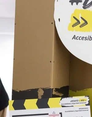 Espacio interior con paneles altos en colores amarillo, negro y marrón que forman un recorrido del proyecto “#Desafío ASU – Accesibilidad y Seguridad Universal”. En primer plano se ve un cartel circular con el nombre del proyecto y, detrás, dos personas observando una estructura del circuito. La escena muestra un entorno expositivo dedicado a la accesibilidad.