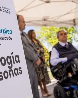 Presentación del proyecto WalkerPisa de Fundación ONCE en un paso de peatones de Las Rozas. En primer plano, un cartel con el lema “Proyecto Walkerpisa. Sensores IoT e IA para pasos de peatones más seguros. Tecnología para las personas”. Al fondo, varias personas durante el acto de presentación, una de ellas utilizando una silla de ruedas, en un entorno urbano al aire libre.