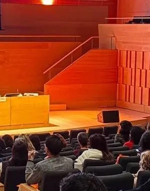 Auditorio moderno con varias decenas de personas sentadas en butacas, asistiendo a una conferencia. En el escenario, una persona realiza una presentación desde un atril mientras otra permanece sentada en una mesa de ponentes. Al fondo se proyecta una diapositiva titulada “Consecuencias del delirium”. La sala cuenta con iluminación cálida, paredes de madera y una amplia zona de gradas ocupada por el público.