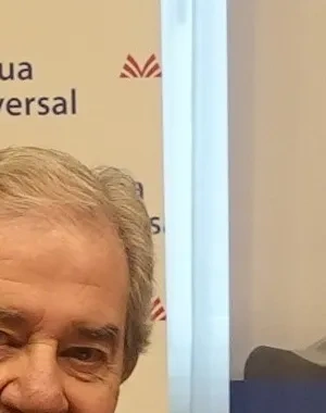 Dos representantes de Mutua Universal y Grupo SIFU estrechan la mano durante la firma de un acuerdo de colaboración. Ambos están sentados frente a una mesa con documentación, junto a paneles corporativos de las dos entidades relacionados con la integración laboral y la inclusión de personas con discapacidad.