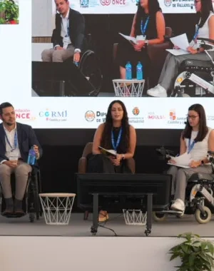 Participantes asisten a una mesa redonda del II Congreso Estatal de Juventud con Discapacidad en Valladolid. En el escenario, tres ponentes intervienen en un debate sobre los 20 años de la Convención de la ONU sobre los Derechos de las Personas con Discapacidad, mientras una intérprete de lengua de signos presta apoyo comunicativo. Al fondo, una gran pantalla reproduce la sesión y, entre el público, se observan asistentes con y sin discapacidad siguiendo la jornada.