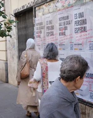 Varias personas caminan y observan una pared cubierta con carteles de una campaña contra el capacitismo en una calle de Madrid. Los carteles muestran frases asociadas a estereotipos sobre la discapacidad, algunas tachadas en rojo, junto a códigos QR y mensajes de sensibilización. En primer plano, una persona se detiene a leer los carteles, mientras al fondo se ve una hilera de árboles y edificios urbanos.
