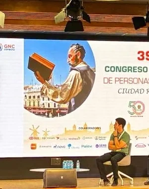 Vista general de un auditorio durante una ponencia del 39º Congreso Nacional de Personas Renales, celebrado en Ciudad Real en 2026. En el escenario, una persona interviene desde un atril mientras varias personas permanecen sentadas en una mesa redonda. Al fondo, una gran pantalla proyecta una presentación sobre el espacio GN Conecta y el papel de “El catalizador”. El público ocupa gran parte de las butacas del auditorio, orientadas hacia el escenario iluminado. A la izquierda se observan las banderas de Esp