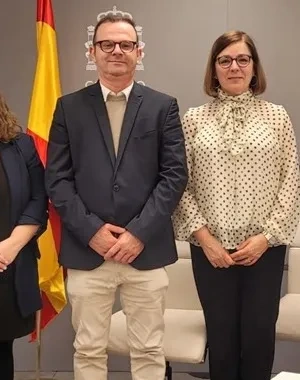 Varias personas posan de pie en una sala institucional frente a un fondo gris con el logotipo del Gobierno de España y dos banderas, una de la Unión Europea y otra de España. Delante hay una mesa con documentos, micrófonos y vasos de agua. El grupo está alineado en el centro del espacio, con sillas blancas en los laterales.