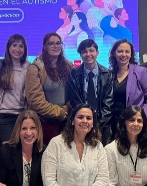 Un grupo numeroso de personas posa en una sala junto a una pantalla que muestra el título “IX Jornada Mujeres en el Autismo. Barreras y buenas prácticas”. La pantalla tiene ilustraciones de varias figuras humanas diversas. A un lado de la imagen hay un panel vertical con el logo de Federación Autismo Madrid repetido. El grupo aparece distribuido en dos filas, algunas personas de pie y otras sentadas, en un espacio interior con sillas transparentes y una mesa baja.
