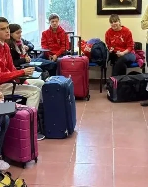 Grupo de jóvenes con sudaderas rojas sentados en un aula o sala de reuniones junto a varias maletas de viaje de distintos colores. En la parte frontal de la sala, varias personas ofrecen indicaciones antes de una salida o actividad organizada. El espacio cuenta con sillas, pupitres, grandes ventanales y equipaje distribuido por la estancia, en un ambiente de preparación para un desplazamiento colectivo.