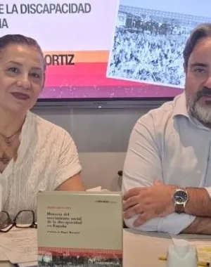 Mesa de presentación de una publicación del CERMI sobre la historia del movimiento social de la discapacidad en España. Dos personas participan en una conferencia junto a ejemplares del libro, micrófonos y material de trabajo. Al fondo, una pantalla muestra la portada de la obra y el título de la presentación virtual.