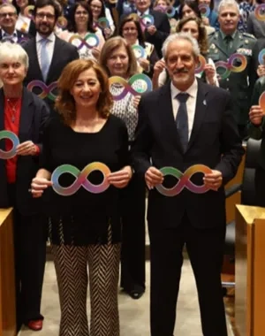 salón de actos del Congreso de los Diputados, lleno de asistentes situados tanto de pie como sentados. Todas las personas sostienen carteles con el símbolo del infinito multicolor, representativo de la diversidad del espectro del autismo. En primera fila, varios participantes permanecen de pie junto a los escaños, mientras el resto del público ocupa el fondo del auditorio. El ambiente es institucional y de apoyo colectivo a la causa del autismo.