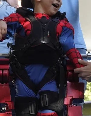 Niño utilizando un exoesqueleto pediátrico asistido por profesionales durante una demostración de neurorrehabilitación. El menor se encuentra sujeto a un dispositivo robótico de marcha sobre una plataforma, mientras personal especializado supervisa y ajusta el equipo ante un público asistente.