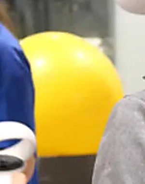 Persona utilizando unas gafas de realidad virtual y mandos de control durante una actividad de rehabilitación tecnológica en un centro especializado. En la sala hay otras dos profesionales con uniforme azul y una pelota terapéutica amarilla al fondo.