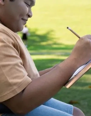 Niño en silla de ruedas escribiendo en un cuaderno a la sombra en un césped