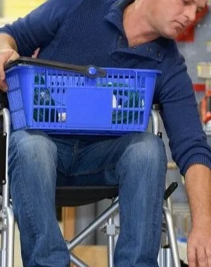 Interior de una tienda de bricolaje o ferretería. Una persona en silla de ruedas se desplaza por un pasillo comercial mientras sostiene una cesta de compra azul sobre las piernas. Con una mano alcanza un producto situado en un estante inferior. A ambos lados del pasillo hay estanterías con herramientas, accesorios y materiales organizados. La imagen muestra una situación de compra y accesibilidad en un entorno comercial.