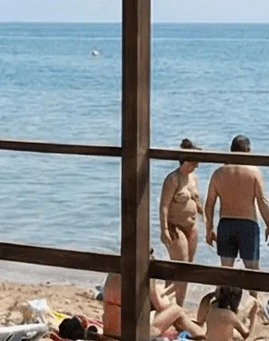 **Alt text:**  Playa con servicio accesible señalizado (“Playa Accesible”) bajo una estructura de madera con pasarela hasta la orilla, donde hay varias sillas anfibias con flotadores naranjas; al fondo, personas en la arena y baño en el mar, y a la derecha una persona usuaria de silla de ruedas acompañada junto al agua.