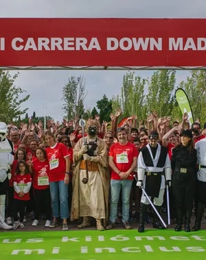 Participantes de la XVI Carrera Down Madrid posan bajo el arco de salida en el Parque Juan Carlos I antes del inicio de la prueba. En primer plano aparecen corredores, voluntarios y personas disfrazadas de personajes de ciencia ficción, mientras una gran multitud se concentra detrás. El arco rojo muestra el nombre del evento y los logotipos de las entidades colaboradoras. La carrera tiene como objetivo promover la inclusión de las personas con discapacidad intelectual a través del deporte.