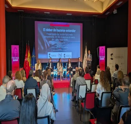 Público asistente y participantes en la presentación del libro Lectura Fácil y web comprensible. Un libro blanco en el marco del nuevo reglamento, celebrada en el Salón de Grados del Campus de Princesa de la Universidad Nebrija. En el escenario, varias personas intervienen en una mesa redonda mientras una persona realiza una presentación desde un atril. Al fondo se proyecta la imagen del evento y se muestran banderas institucionales.