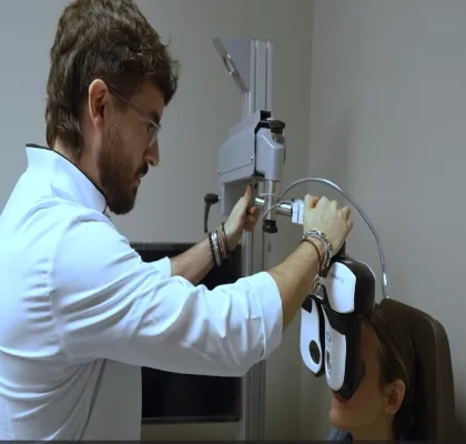 Profesional sanitario realiza una prueba de visión a una persona sentada utilizando un equipo optométrico en una consulta. La persona examinada lleva colocado un dispositivo frente a los ojos mientras el especialista ajusta el aparato para evaluar su capacidad visual. El entorno es una sala clínica con equipamiento oftalmológico.