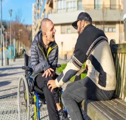 Dos hombres, un de ellos en silla de ruedas y el otro sentado en un banco, hablando