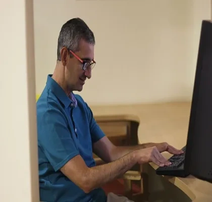 Persona sentada frente a un ordenador de sobremesa, utilizando el teclado en un entorno de trabajo interior. La imagen muestra un puesto de oficina con una pantalla, una mesa de madera y una iluminación suave.