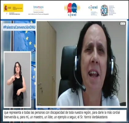 Captura de una videoconferencia institucional. En el centro, una persona sentada en un despacho, frente a la cámara, con auriculares y micrófono, interviene desde su puesto de trabajo. A la izquierda de la pantalla aparece una intérprete de lengua de signos española sobre fondo neutro. En el margen superior se muestran los logotipos del Gobierno de España, Fundación CERMI Mujeres y otras entidades, junto al texto “#PalestraConvenciónONU”. En la parte inferior se ven subtítulos en español.