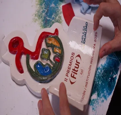 Trofeo de cerámica roto con diseño colorido y texto de los "II PREMIOS Fitur 4all 2025". Manos lo sostienen sobre una mesa con pintura, sugiriendo reparación o restauración.