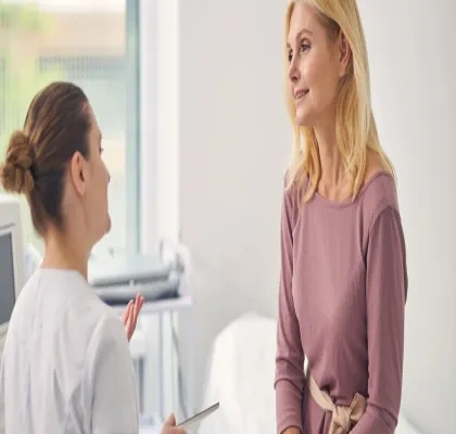 Persona sentada en una consulta médica conversa con un profesional sanitario. Ambos aparecen en una sala de exploración luminosa, con equipamiento clínico al fondo, en una escena relacionada con el asesoramiento y seguimiento de la salud durante la menopausia.