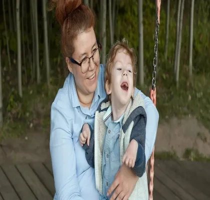 Persona adulta con gafas sostiene a un niño con discapacidad en un columpio adaptado al aire libre. Ambos se encuentran en una zona de juego rodeada de árboles. La imagen refleja acompañamiento, inclusión y apoyo a la infancia con discapacidad en un entorno accesible y seguro.