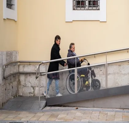 hombre ayudando a una persona en silla de ruedas subir por una rampa accediendo a un edificio.