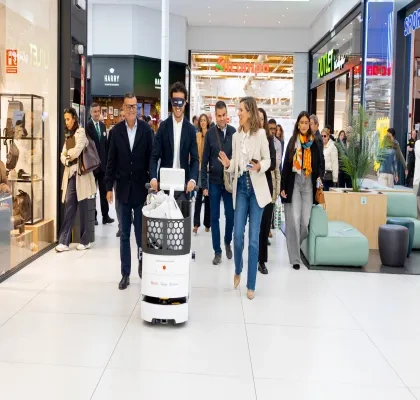 Persona guiando un robot asistencial autónomo por el pasillo de un centro comercial. Varias personas acompañan la demostración y observan el recorrido del dispositivo, en un espacio comercial amplio y luminoso con tiendas y zonas de descanso al fondo. La imagen muestra una prueba de tecnología orientada a mejorar la accesibilidad y la movilidad en entornos comerciales.