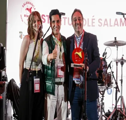 Tres personas posan sobre un escenario durante un acto de Startup Olé Salamanca. Una de ellas sostiene un premio de color rojo mientras otra hace el gesto de aprobación con el pulgar. En primer plano hay varios micrófonos y, al fondo, se aprecia una batería y el logotipo del evento proyectado en una pantalla.