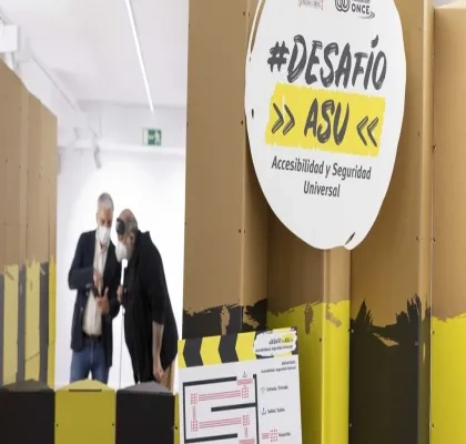 Espacio interior con paneles altos en colores amarillo, negro y marrón que forman un recorrido del proyecto “#Desafío ASU – Accesibilidad y Seguridad Universal”. En primer plano se ve un cartel circular con el nombre del proyecto y, detrás, dos personas observando una estructura del circuito. La escena muestra un entorno expositivo dedicado a la accesibilidad.