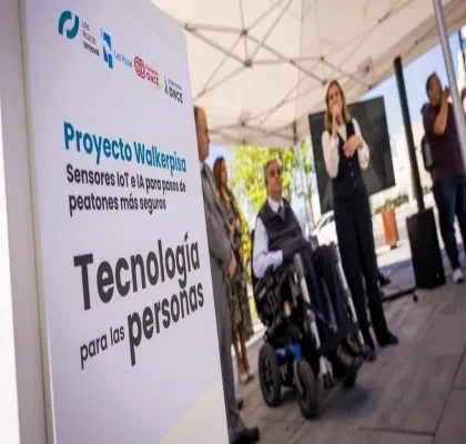 Presentación del proyecto WalkerPisa de Fundación ONCE en un paso de peatones de Las Rozas. En primer plano, un cartel con el lema “Proyecto Walkerpisa. Sensores IoT e IA para pasos de peatones más seguros. Tecnología para las personas”. Al fondo, varias personas durante el acto de presentación, una de ellas utilizando una silla de ruedas, en un entorno urbano al aire libre.