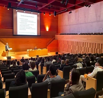Auditorio moderno con varias decenas de personas sentadas en butacas, asistiendo a una conferencia. En el escenario, una persona realiza una presentación desde un atril mientras otra permanece sentada en una mesa de ponentes. Al fondo se proyecta una diapositiva titulada “Consecuencias del delirium”. La sala cuenta con iluminación cálida, paredes de madera y una amplia zona de gradas ocupada por el público.