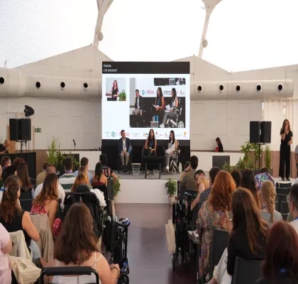 Participantes asisten a una mesa redonda del II Congreso Estatal de Juventud con Discapacidad en Valladolid. En el escenario, tres ponentes intervienen en un debate sobre los 20 años de la Convención de la ONU sobre los Derechos de las Personas con Discapacidad, mientras una intérprete de lengua de signos presta apoyo comunicativo. Al fondo, una gran pantalla reproduce la sesión y, entre el público, se observan asistentes con y sin discapacidad siguiendo la jornada.
