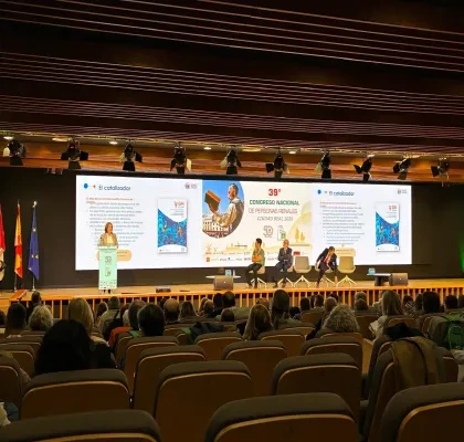 Vista general de un auditorio durante una ponencia del 39º Congreso Nacional de Personas Renales, celebrado en Ciudad Real en 2026. En el escenario, una persona interviene desde un atril mientras varias personas permanecen sentadas en una mesa redonda. Al fondo, una gran pantalla proyecta una presentación sobre el espacio GN Conecta y el papel de “El catalizador”. El público ocupa gran parte de las butacas del auditorio, orientadas hacia el escenario iluminado. A la izquierda se observan las banderas de Esp