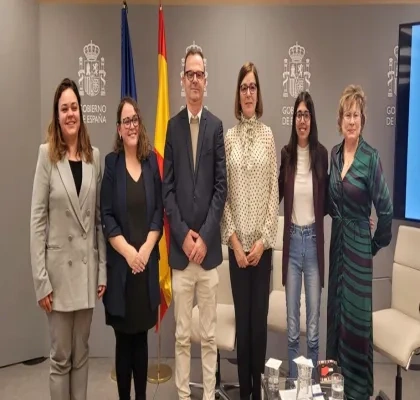 Varias personas posan de pie en una sala institucional frente a un fondo gris con el logotipo del Gobierno de España y dos banderas, una de la Unión Europea y otra de España. Delante hay una mesa con documentos, micrófonos y vasos de agua. El grupo está alineado en el centro del espacio, con sillas blancas en los laterales.