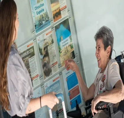 Una dependienta de la agencia de viajes enseñando folletos a una señora en silla de ruedas