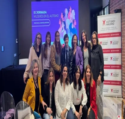 Un grupo numeroso de personas posa en una sala junto a una pantalla que muestra el título “IX Jornada Mujeres en el Autismo. Barreras y buenas prácticas”. La pantalla tiene ilustraciones de varias figuras humanas diversas. A un lado de la imagen hay un panel vertical con el logo de Federación Autismo Madrid repetido. El grupo aparece distribuido en dos filas, algunas personas de pie y otras sentadas, en un espacio interior con sillas transparentes y una mesa baja.