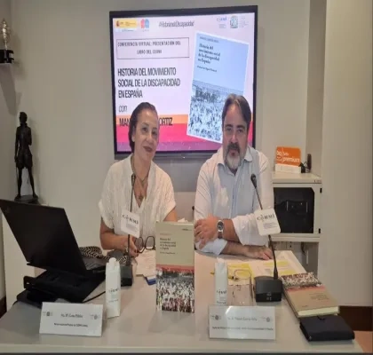 Mesa de presentación de una publicación del CERMI sobre la historia del movimiento social de la discapacidad en España. Dos personas participan en una conferencia junto a ejemplares del libro, micrófonos y material de trabajo. Al fondo, una pantalla muestra la portada de la obra y el título de la presentación virtual.