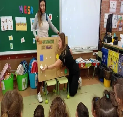 Escena en un aula infantil con dos maestras y un grupo de niños sentados en el suelo. Una maestra, vestida con una blusa gris y jeans, está de pie, mientras la otra, con ropa negra, interactúa con un modelo grande de mesa de madera con taburetes y un cartel que dice 'POR 4 ESQUINITAS DE NADA'. Fondo con pizarrón verde, dibujos, letras y materiales escolares. Ambiente colorido y educativo