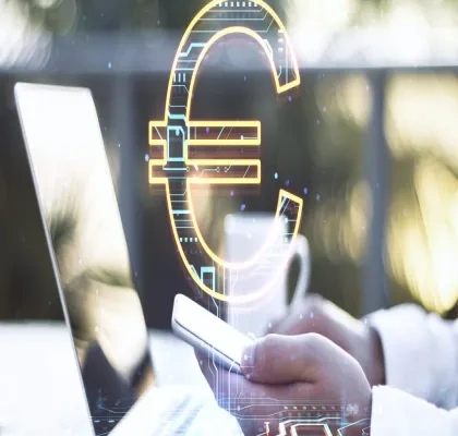 Mano sosteniendo un teléfono móvil junto a un ordenador portátil, con un símbolo del euro digital superpuesto en el centro de la imagen. La escena representa conceptos relacionados con pagos electrónicos, finanzas digitales y tecnología financiera. Fondo desenfocado con tonos claros y elementos tecnológicos.