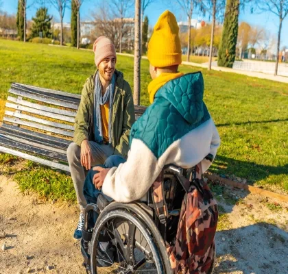 Persona en silla de ruedas y otra persona mantienen una conversación en un parque urbano con zonas verdes y caminos peatonales.