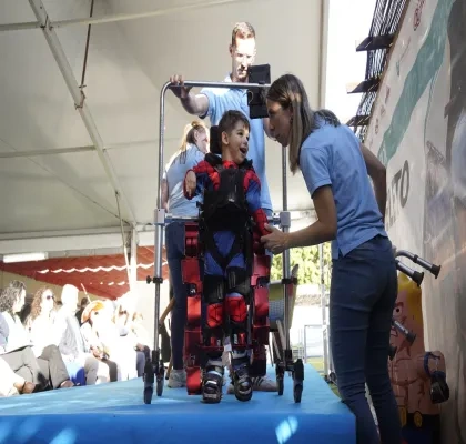 Niño utilizando un exoesqueleto pediátrico asistido por profesionales durante una demostración de neurorrehabilitación. El menor se encuentra sujeto a un dispositivo robótico de marcha sobre una plataforma, mientras personal especializado supervisa y ajusta el equipo ante un público asistente.