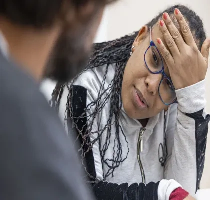 Persona con gafas conversa con otra persona en un entorno interior. Apoya la cabeza sobre una mano mientras escucha, en una escena relacionada con atención, apoyo o acompañamiento profesional.