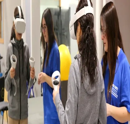 Persona utilizando unas gafas de realidad virtual y mandos de control durante una actividad de rehabilitación tecnológica en un centro especializado. En la sala hay otras dos profesionales con uniforme azul y una pelota terapéutica amarilla al fondo.