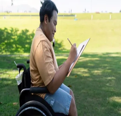 Niño en silla de ruedas escribiendo en un cuaderno a la sombra en un césped