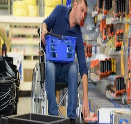 Interior de una tienda de bricolaje o ferretería. Una persona en silla de ruedas se desplaza por un pasillo comercial mientras sostiene una cesta de compra azul sobre las piernas. Con una mano alcanza un producto situado en un estante inferior. A ambos lados del pasillo hay estanterías con herramientas, accesorios y materiales organizados. La imagen muestra una situación de compra y accesibilidad en un entorno comercial.