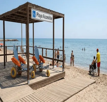 **Alt text:**  Playa con servicio accesible señalizado (“Playa Accesible”) bajo una estructura de madera con pasarela hasta la orilla, donde hay varias sillas anfibias con flotadores naranjas; al fondo, personas en la arena y baño en el mar, y a la derecha una persona usuaria de silla de ruedas acompañada junto al agua.