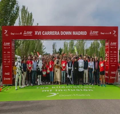 Participantes de la XVI Carrera Down Madrid posan bajo el arco de salida en el Parque Juan Carlos I antes del inicio de la prueba. En primer plano aparecen corredores, voluntarios y personas disfrazadas de personajes de ciencia ficción, mientras una gran multitud se concentra detrás. El arco rojo muestra el nombre del evento y los logotipos de las entidades colaboradoras. La carrera tiene como objetivo promover la inclusión de las personas con discapacidad intelectual a través del deporte.