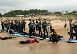 Participantes y monitores de una actividad de surf adaptado reciben formación en la playa de Loredo durante el I Festival de Parasurf Fundación También–Plea. Varias personas practican la posición sobre tablas de surf en la arena mientras un grupo observa y se prepara para entrar al agua. La imagen muestra una jornada de deporte inclusivo y aprendizaje junto al mar Cantábrico.