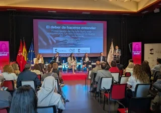 Público asistente y participantes en la presentación del libro Lectura Fácil y web comprensible. Un libro blanco en el marco del nuevo reglamento, celebrada en el Salón de Grados del Campus de Princesa de la Universidad Nebrija. En el escenario, varias personas intervienen en una mesa redonda mientras una persona realiza una presentación desde un atril. Al fondo se proyecta la imagen del evento y se muestran banderas institucionales.